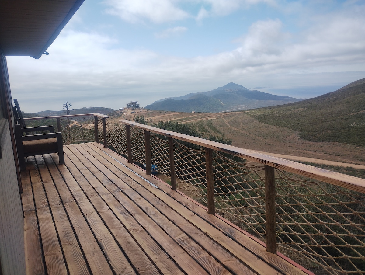 A wooden deck is shown with railing, overlooking a scenic view of rolling hills and distant mountains. The deck features a pair of sturdy chairs, and clouds drift across a clear sky, creating a serene environment.