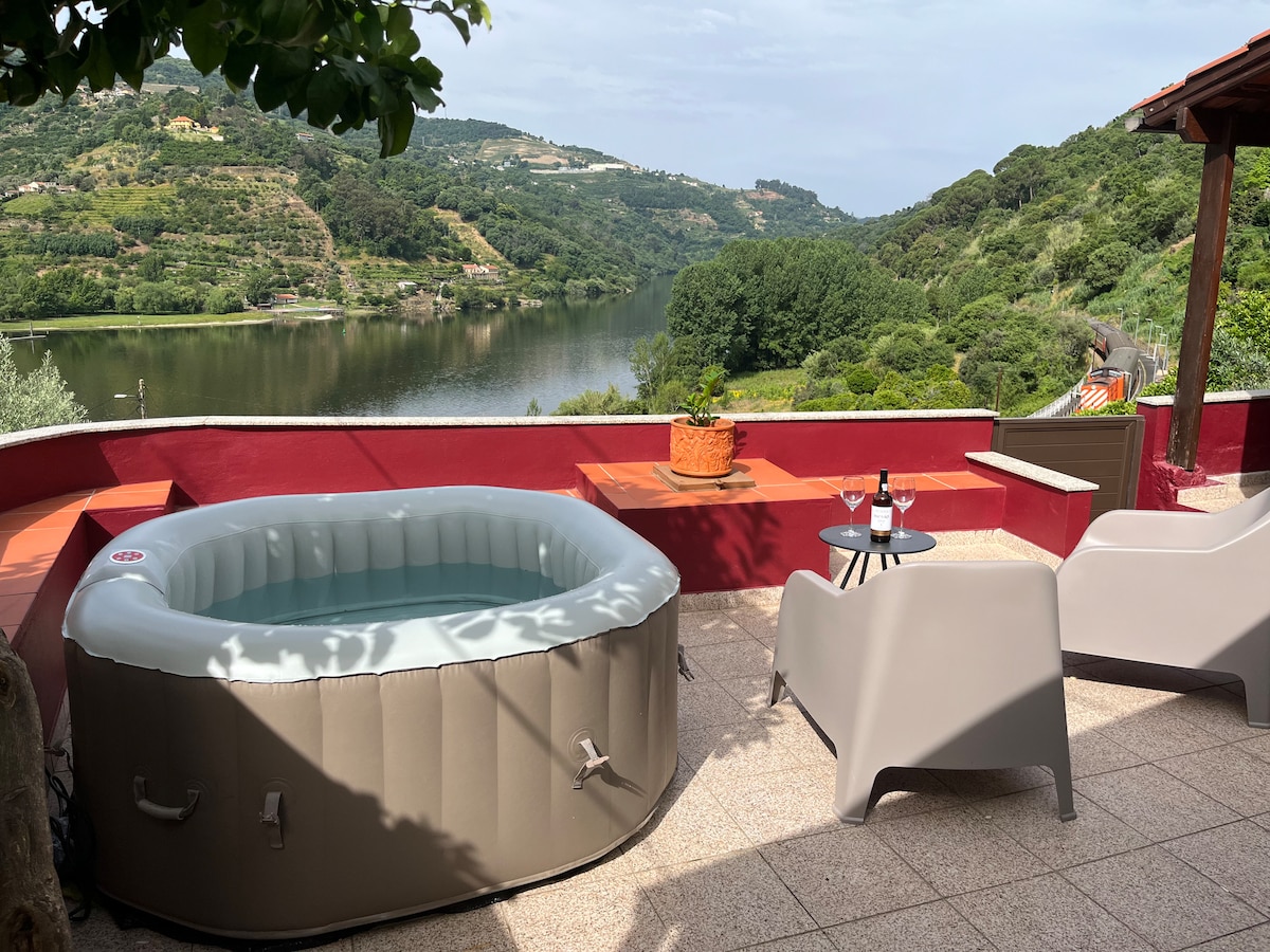 An outdoor terrace features a round inflatable jacuzzi, surrounded by greenery and offering views of the Douro River. Two modern chairs and a small table with wine bottles sit nearby, inviting relaxation in a serene setting.