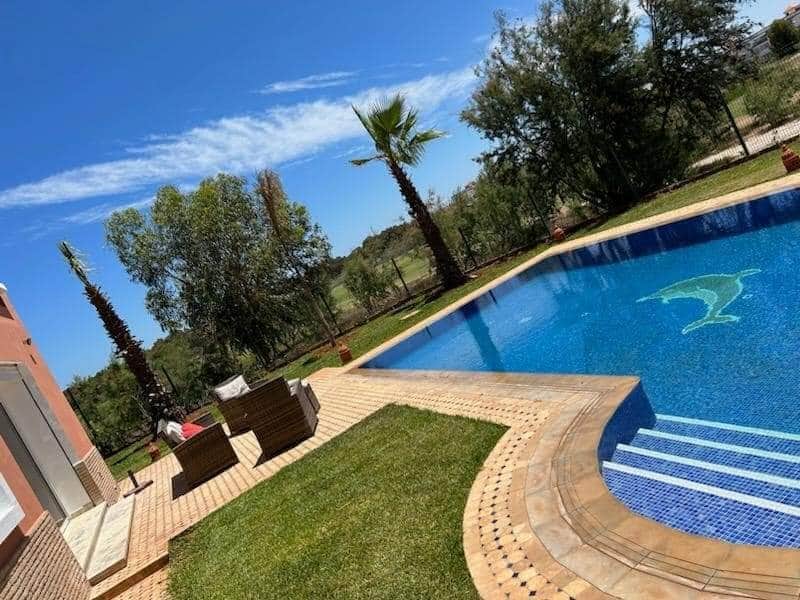 An outdoor area features a shimmering swimming pool with a dolphin design at the bottom. Lush greenery surrounds the space, complimented by palm trees. Comfortable seating is positioned on a patio beside the pool, with a clear blue sky overhead.