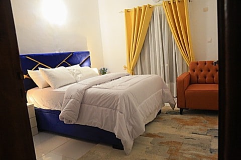 A cozy bedroom is presented with a spacious bed adorned with white bedding and decorative pillows. A vibrant blue headboard contrasts with the soft yellow curtains framing the window. A comfortable orange armchair adds a pop of color to the room's inviting ambiance.