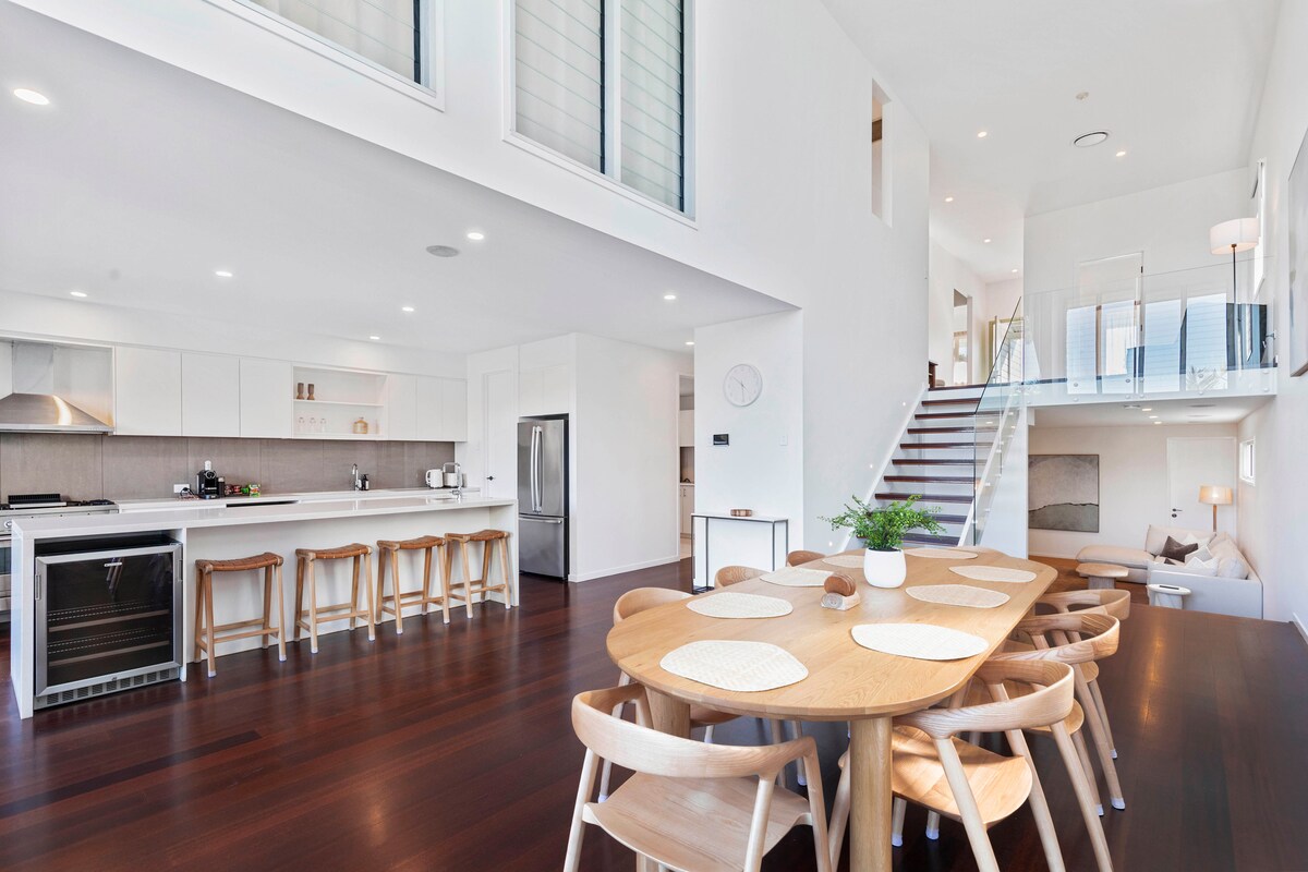 A bright open-plan living and dining area features a modern kitchen with sleek cabinetry and stainless steel appliances. A wooden dining table is surrounded by light chairs, and a staircase leads to an upper level. Large windows allow natural light to illuminate the space.