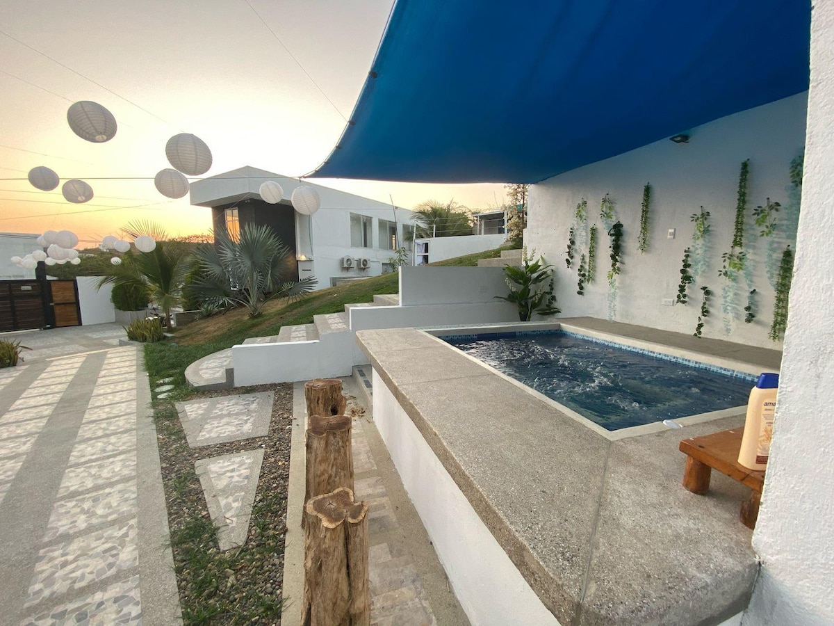 An outdoor area features a small hot tub nestled beside a tree-lined pathway. Soft ambient lighting from hanging lanterns enhances the surroundings, while a gentle slope leads to a modern building in the background, partially shaded by a blue canopy.