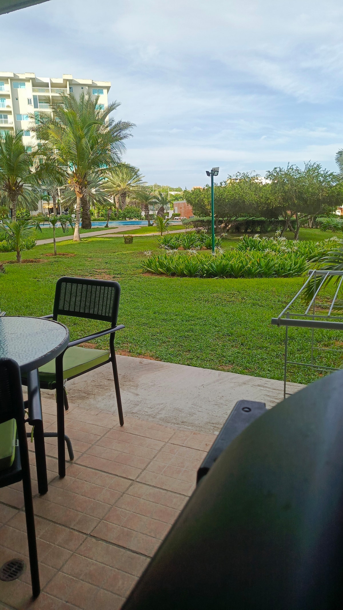 A serene outdoor terrace is shown, featuring a round table and four chairs. Lush greenery surrounds the area, including palm trees and manicured lawns, creating a peaceful view of the garden. The space allows for outdoor dining and relaxation.