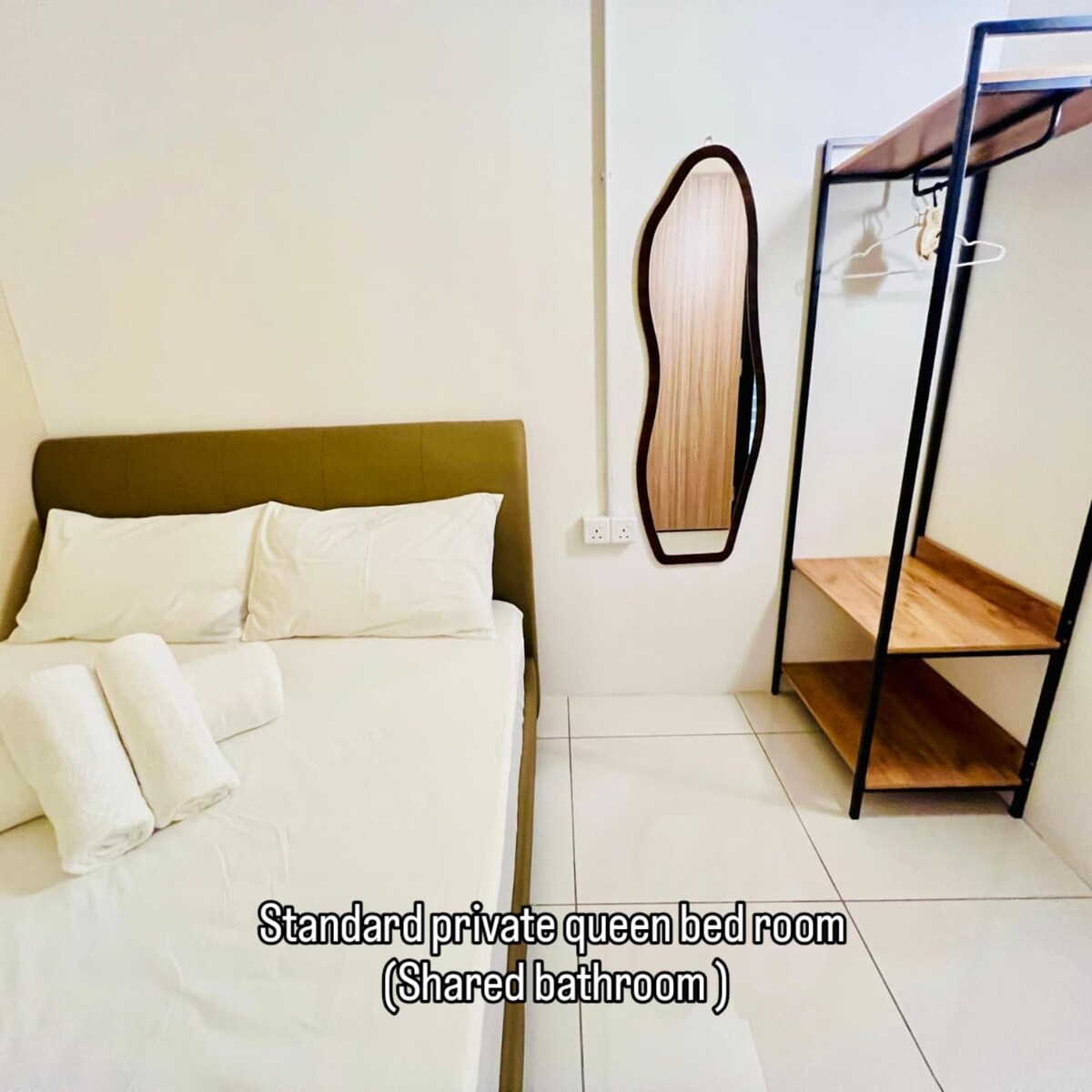 A standard private room is presented with a queen-sized bed adorned with soft white linens and neatly arranged towels. An elegant mirror complements the room, while a metal frame with wooden shelves offers storage options. The minimalist design features light-colored walls and tiled flooring.