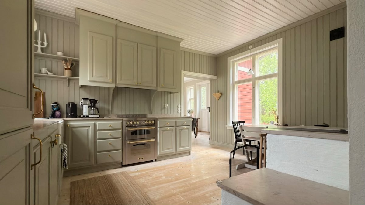 A modern kitchen is presented with grey cabinets and stainless steel appliances. A large oven is positioned beneath a decorative hood. A small dining area is visible, featuring a black chair next to a white table. Light enters through two windows, illuminating the wooden floor.