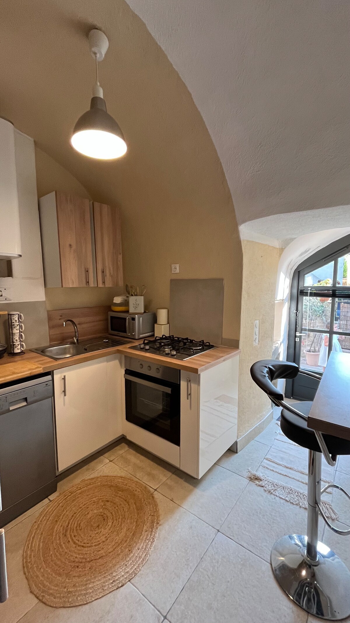 A compact kitchen is presented with a blend of light wood and white cabinetry. A stovetop and oven are positioned under a muted archway, while a small breakfast bar with two black stools is situated near a window. A natural fiber rug adds texture to the tile flooring.