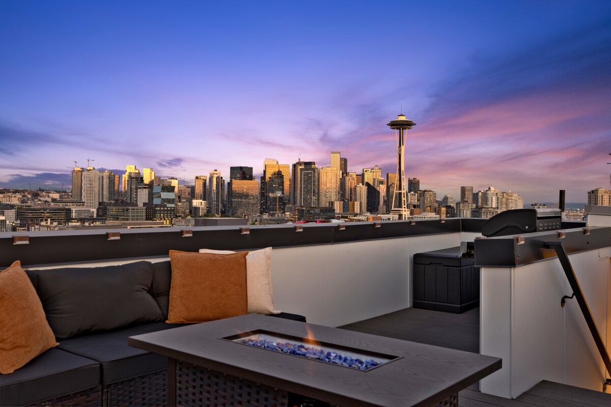 The rooftop deck offers expansive views of the Seattle skyline, highlighted by the Space Needle. A comfortable seating arrangement is positioned near a gas fire table, with inviting pillows enhancing the space. A BBQ grill is visible, ready for outdoor cooking.