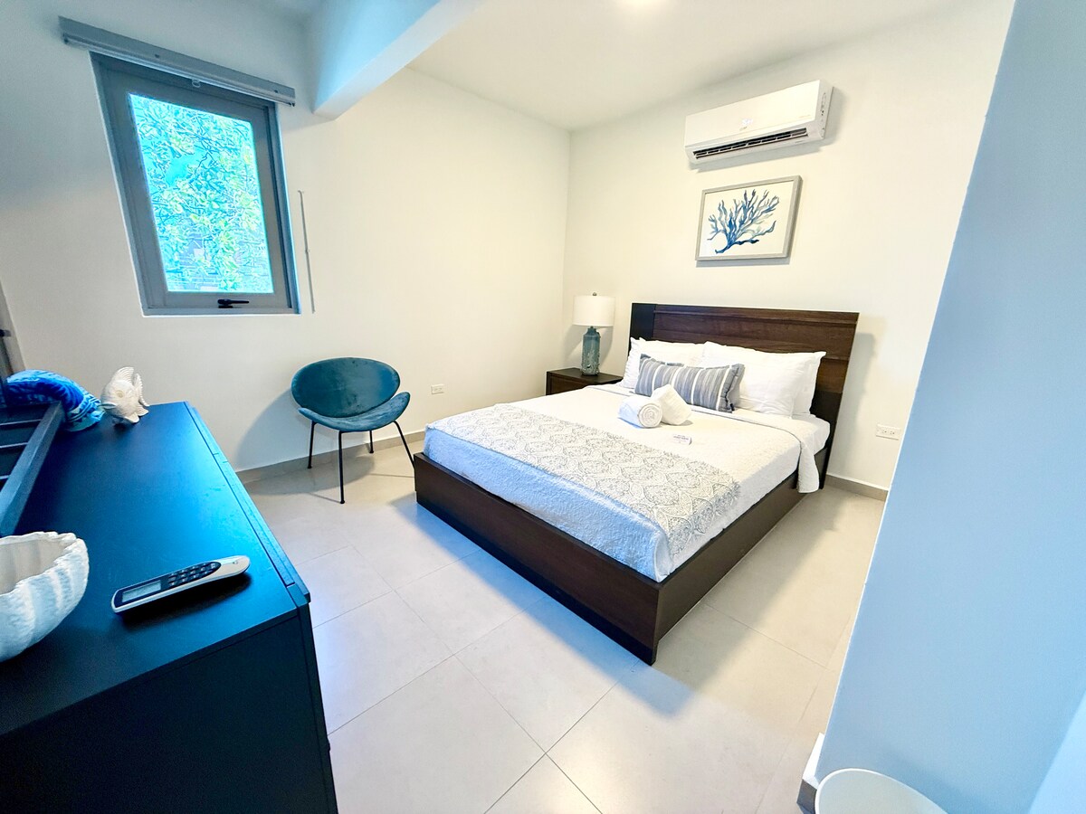 Acogedor y luminoso dormitorio con una cama tamaño queen, sábanas suaves, aire acondicionado y muebles modernos. Incluye una cómoda con espejo, una cómoda silla decorativa y un vestidor con perchas y estantes de almacenamiento para tu comodidad.