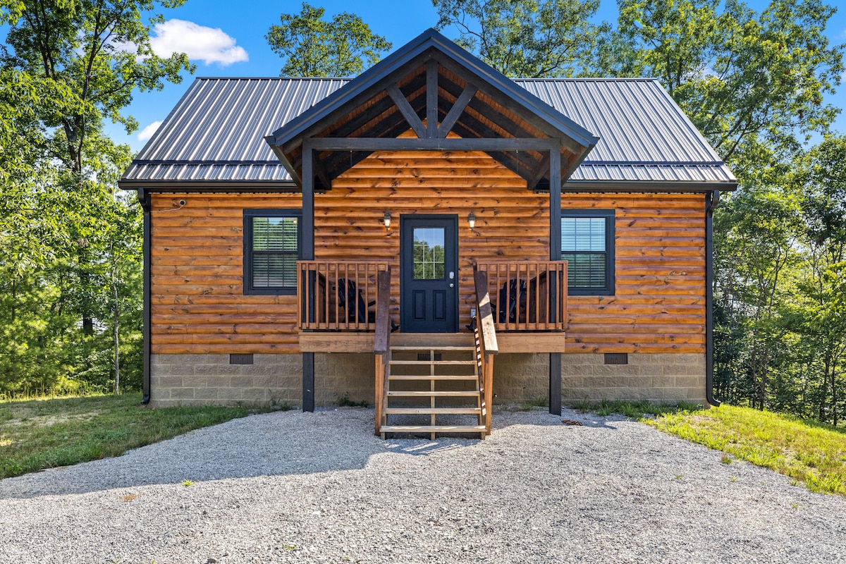 The cabin features a rustic wooden exterior with a steep, metal roof. A set of stairs leads to the front door, flanked by two windows. Surrounding the cabin are trees and a gravel path, enhancing the natural setting of the location.