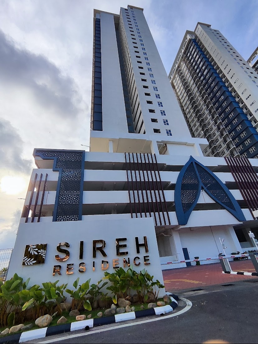 The exterior view of Sireh Residence is showcased, featuring a modern building structure with multiple floors. A prominent sign displays the name 'SIREH RESIDENCE.' Lush greenery surrounds the entrance, and the sky is partially cloudy, enhancing the building's contemporary architecture.