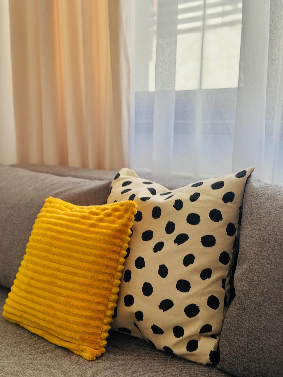 A cozy seating area features two decorative cushions on a soft gray sofa. One cushion is textured with deep yellow stripes, while the other displays a pattern of black dots on a light background. Natural light filters through sheer curtains, creating a soft ambiance.