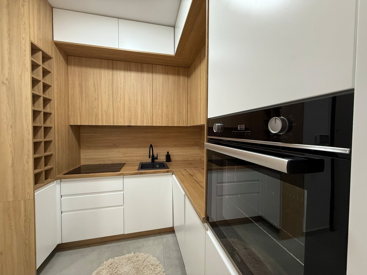 A modern kitchen is displayed with a combination of sleek white cabinetry and warm wooden accents. An integrated oven is positioned at eye level, while a minimalist sink is situated on a matching countertop. Soft textures are introduced by a round rug on the concrete floor.