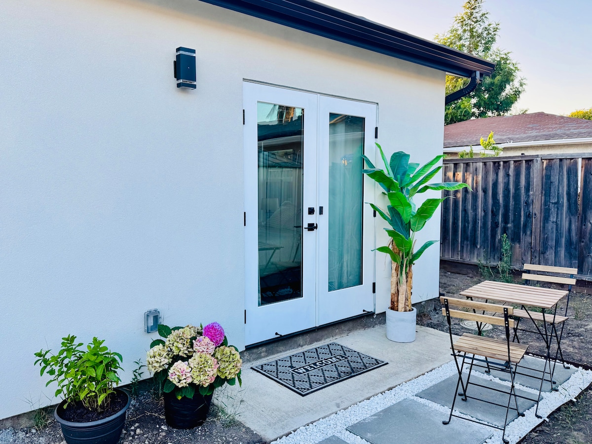 The exterior entrance features double glass doors inviting guests into the suite. A small patio area contains a bistro table with two chairs, complemented by potted plants that add a touch of greenery. Pebbled pathways lead to the entrance, enhancing the welcoming appearance.