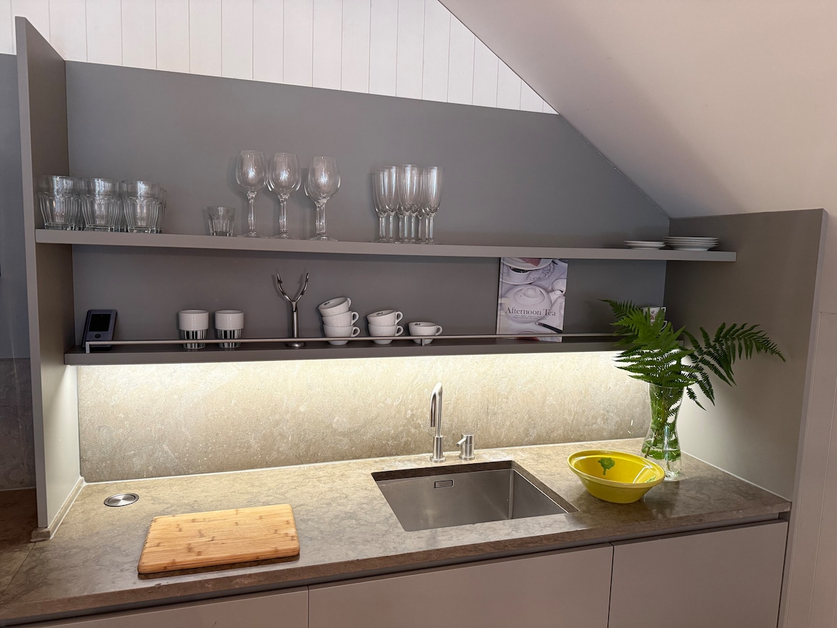 A modern kitchen area is showcased with sleek cabinetry and a minimalist design. Glassware is neatly arranged on floating shelves above a stone countertop. A sink is built into the countertop, accompanied by a cutting board and a bright yellow bowl, enhancing the functional space.