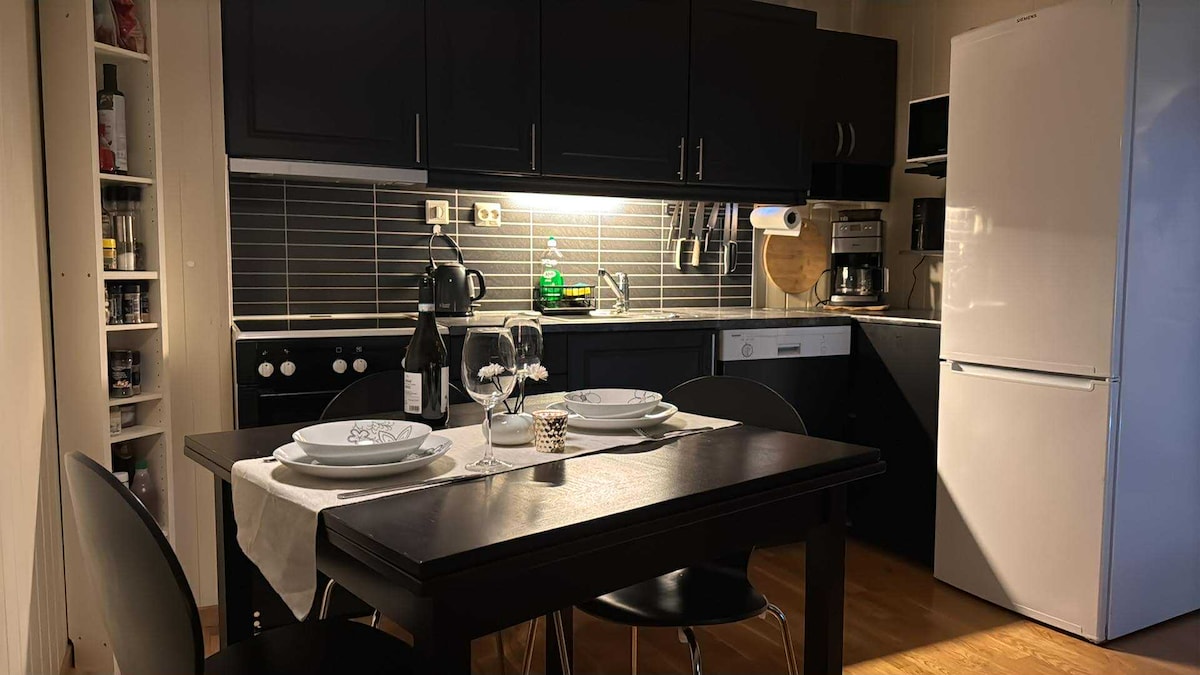 A compact kitchen area features dark cabinetry and modern appliances. A dining table is set for two, complete with plates, glasses, and a decorative centerpiece. Bright overhead lighting highlights the sleek countertops and tiled backsplash, lending an inviting ambiance to the space.