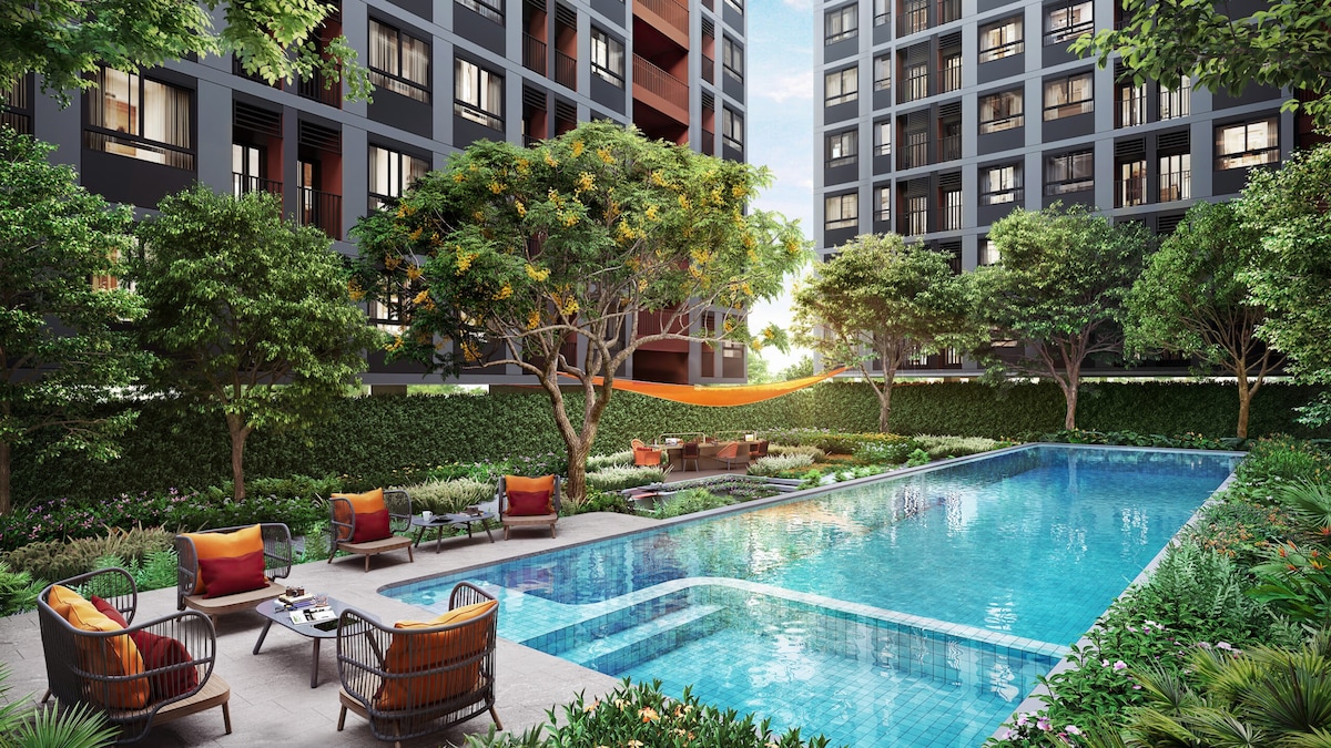 A modern pool area is surrounded by well-maintained greenery and trees, offering a tranquil environment. Lounge chairs with cushions and a hammock provide relaxation options, while the swimming pool reflects the sunlight. Nearby buildings feature large windows that enhance the inviting atmosphere.