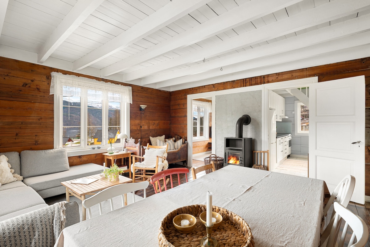 A bright and airy living space showcases wooden beams and a cozy atmosphere. A dining table is set in the foreground, while a comfortable seating area includes soft couches and decorative cushions. A modern stove is positioned in the corner, enhancing the inviting environment.