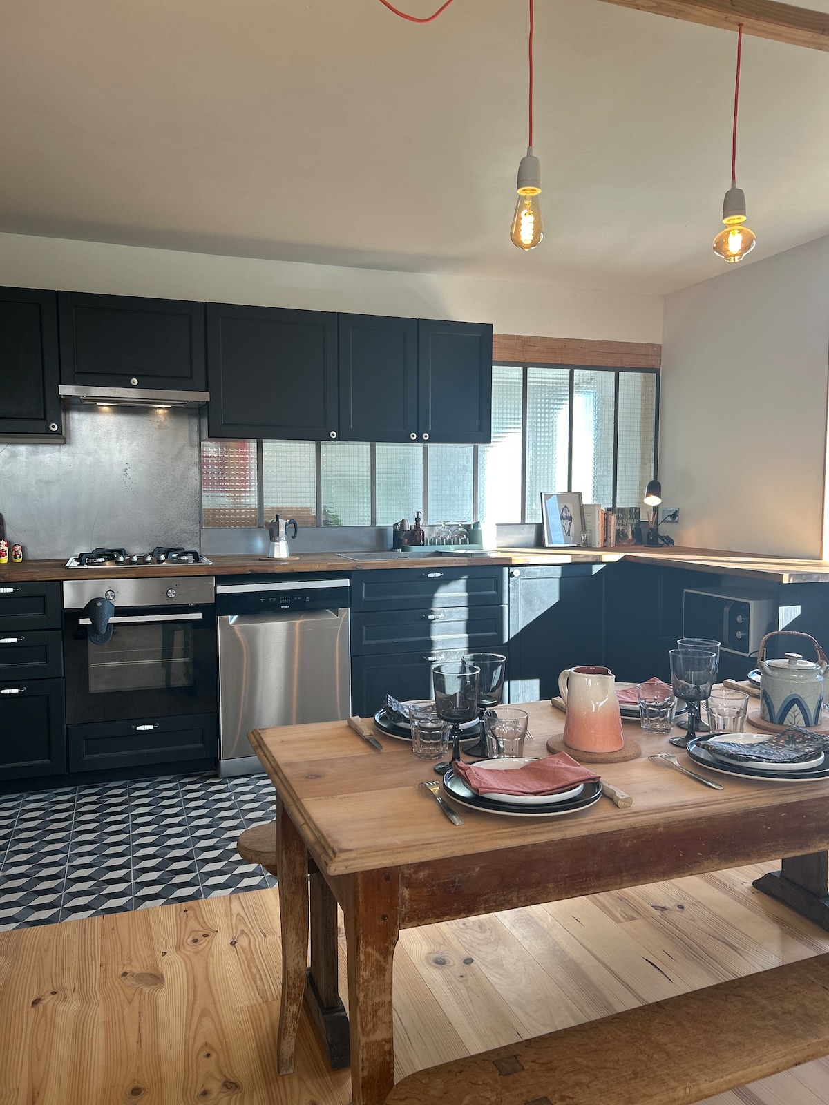 The kitchen features dark cabinetry with modern appliances and a polished countertop. A dining table is set for a meal with plates, glasses, and a central pitcher. Natural light filters through a large window, illuminating the space and showcasing the patterned floor.