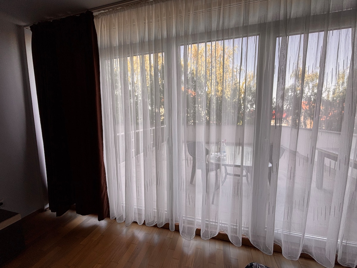 Floor-to-ceiling sheer curtains softly filter natural light, illuminating the spacious indoor area. Outside, a private balcony is visible, featuring simple outdoor furniture that invites relaxation. The view is framed by tree foliage, enhancing the sense of tranquility.