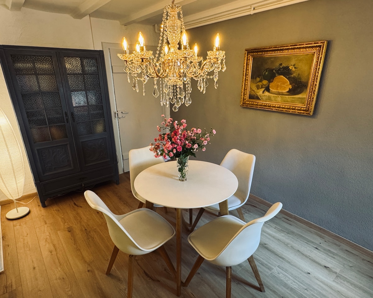 A dining area features a round table surrounded by four light-colored chairs. Above, a crystal chandelier casts a soft glow, while a vase of fresh flowers adds a touch of color. A decorative painting hangs on the wall, complementing the overall aesthetic.