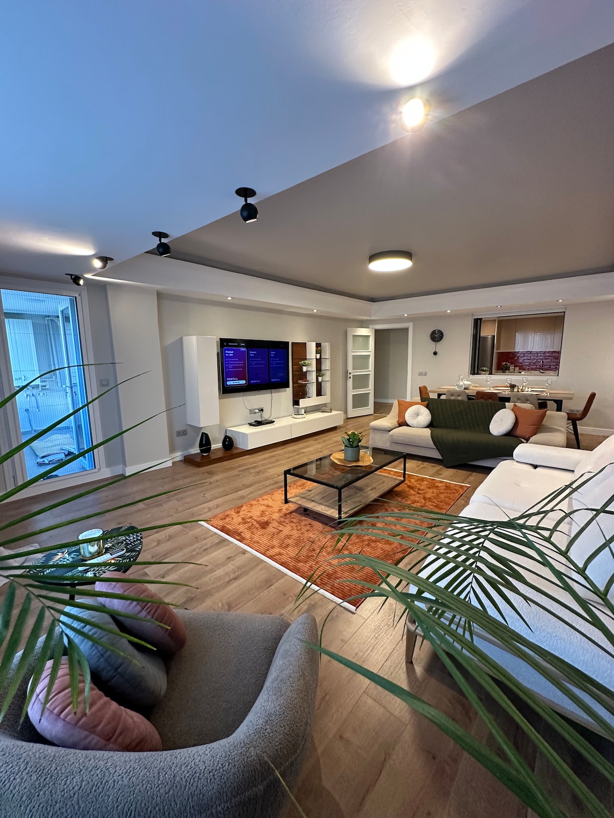 A modern living area is highlighted by a spacious white sofa, positioned across from a wall-mounted TV. A coffee table sits center stage, adorned with potted greenery. Natural light filters through large windows, while a textured area rug defines the seating space.