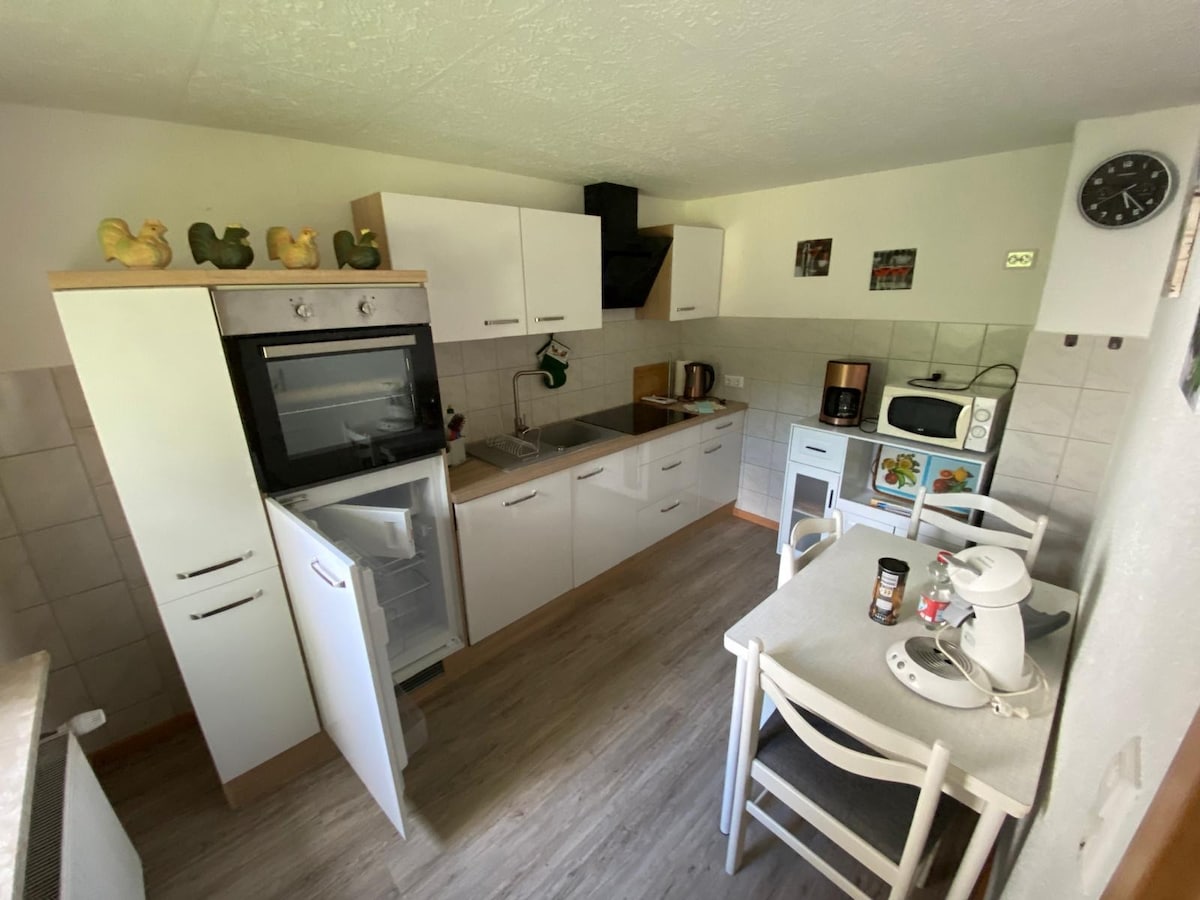 A modern kitchen is presented with white cabinetry and stainless steel appliances. A refrigerator and an oven are installed, alongside a microwave and a coffee maker. A small dining table with four chairs is positioned against one wall, enhancing the space's functionality.