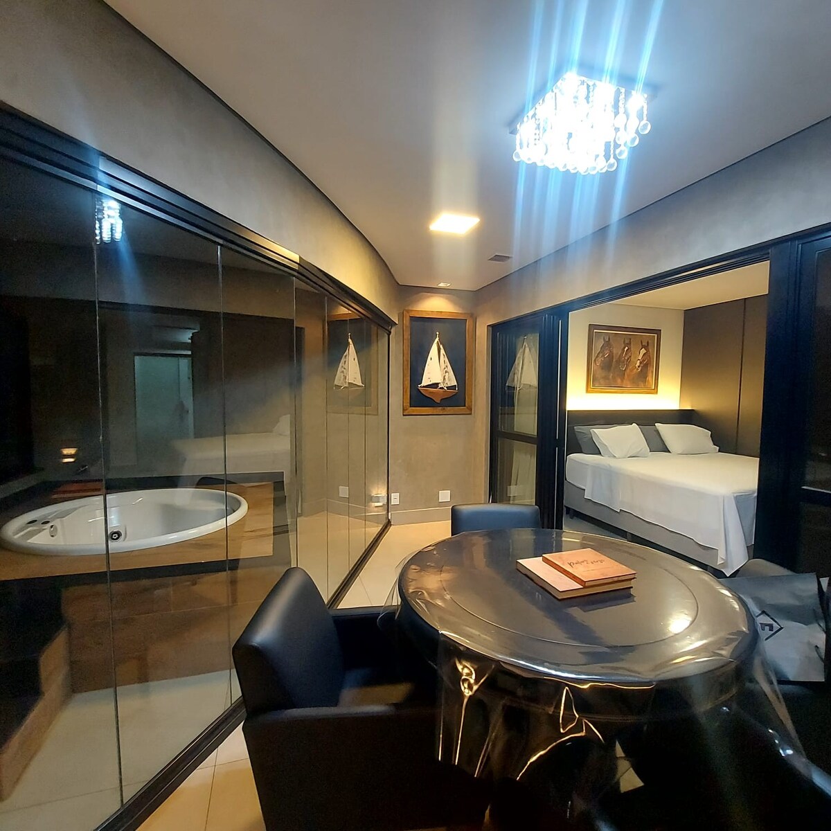 A modern and stylish interior features a glass-walled jacuzzi area, a cozy sleeping area with a spacious bed, and a dining space with a round table and black chairs. Soft lighting from a chandelier enhances the atmosphere.