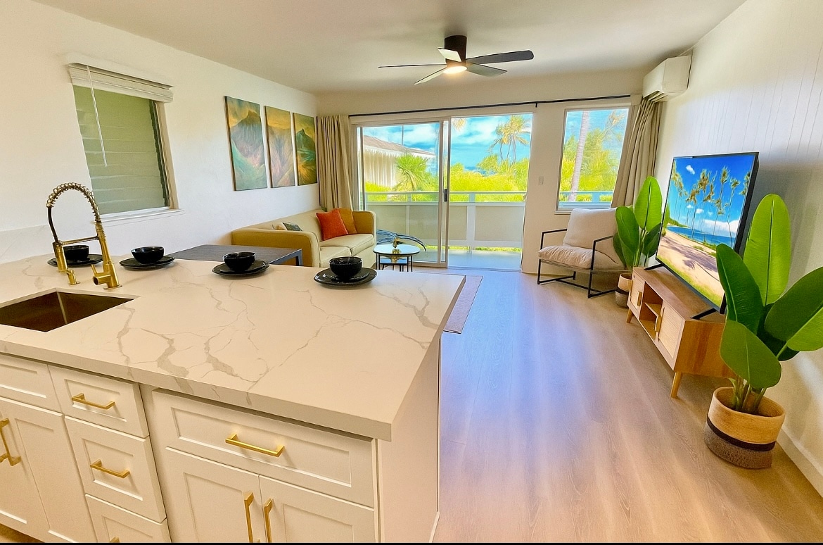 A modern living area features a comfortable sofa and a light-colored armchair, with large windows allowing natural light. A sleek kitchen island with marble countertops is visible in the foreground, and tropical plants add a touch of greenery. Neutral tones create a serene ambiance.