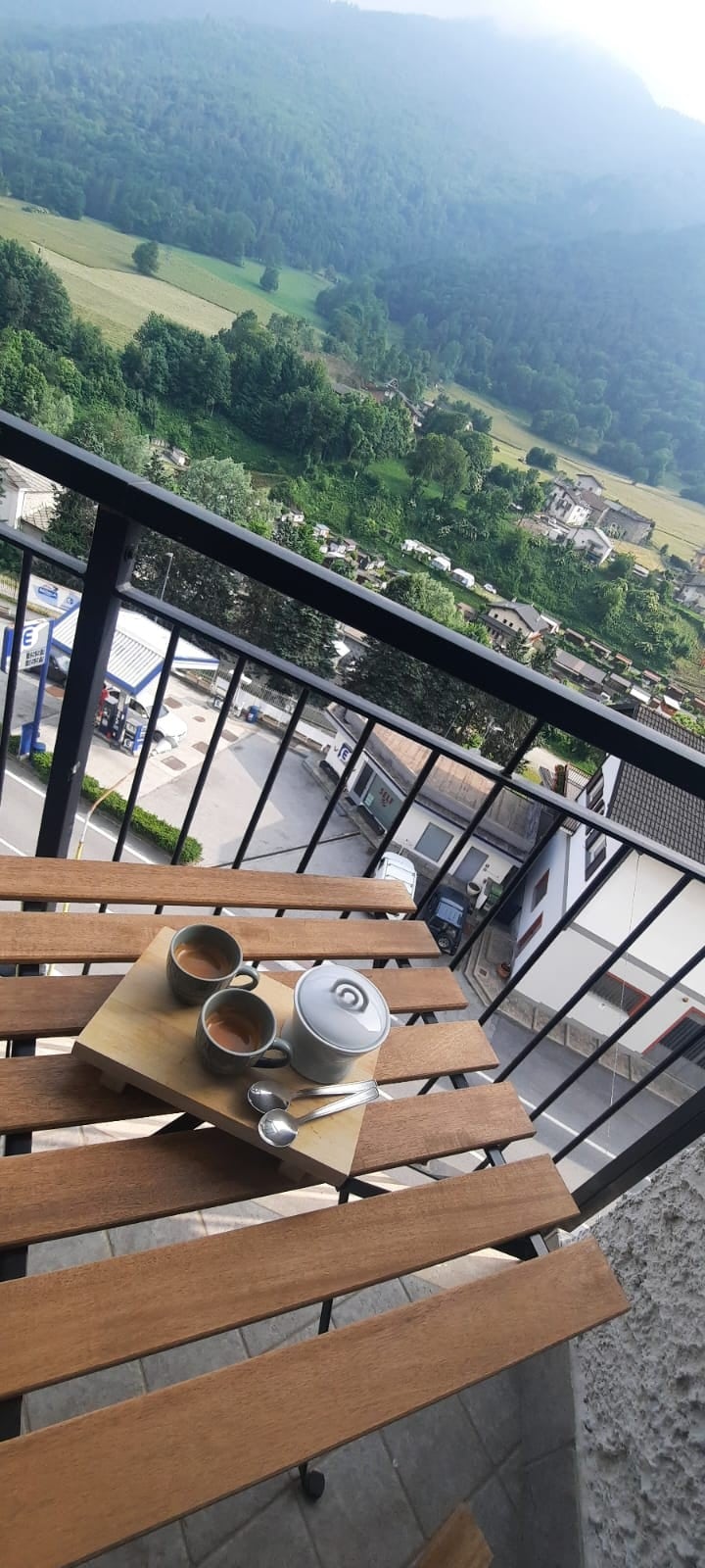A wooden table is set on a balcony, featuring a teapot and two cups. An expansive view of green hills and distant buildings can be seen beyond the railing, enhancing the tranquil atmosphere of the outdoor space.