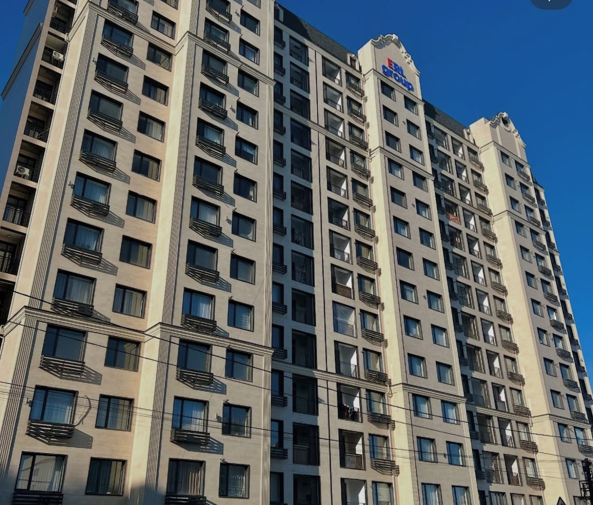 A modern building is presented with a light-colored facade and a symmetrical arrangement of windows. Balconies are visible on several floors, contributing to the structure's design. The clear blue sky serves as a backdrop, accentuating the building's contemporary style.