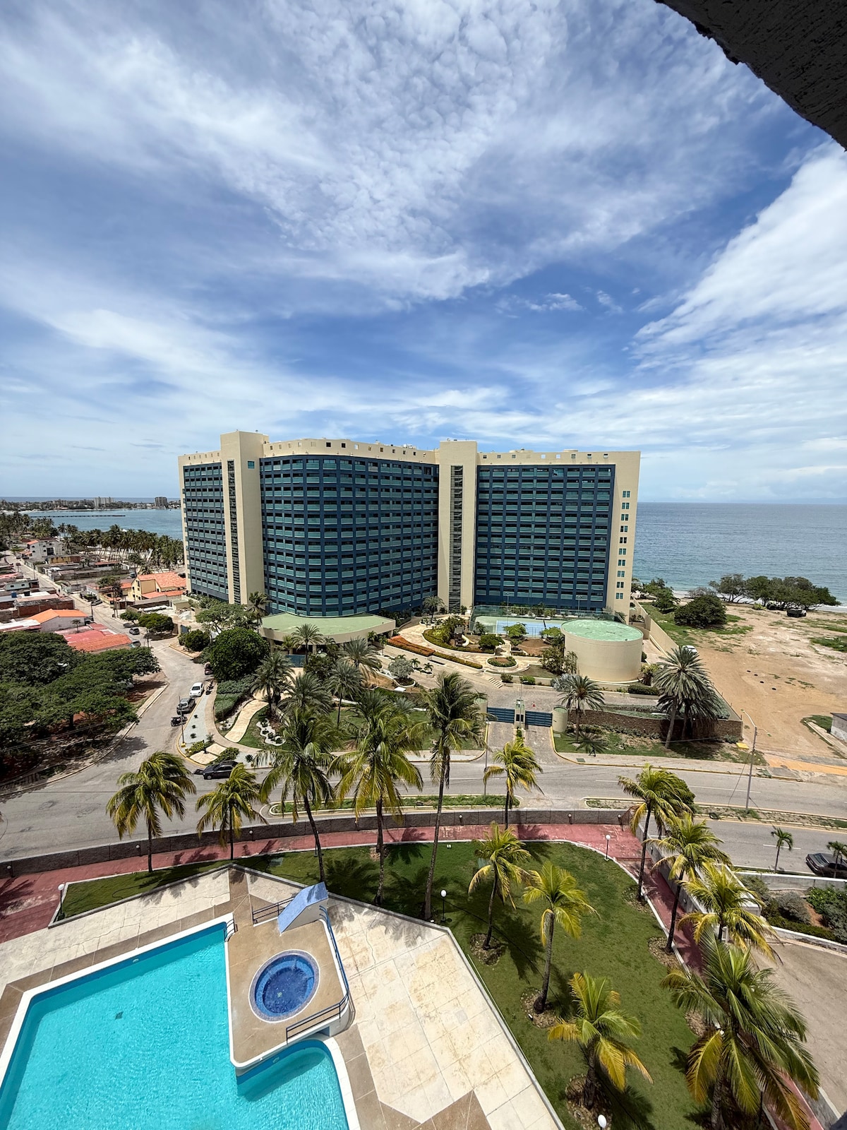 An aerial view captures a modern building near the coastline, highlighted by a clear blue sky and scattered clouds. A shimmering swimming pool is visible in the foreground, surrounded by a well-maintained area with palm trees, contributing to a relaxing outdoor environment.