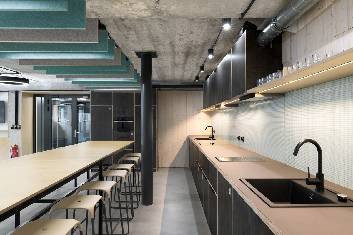 A modern kitchen is displayed featuring minimalist cabinetry and a long wooden countertop. Sleek black fixtures and a large sink are positioned beneath ambient lighting. Barstools are arranged along one side of the countertop, with a textured ceiling adding visual interest overhead.