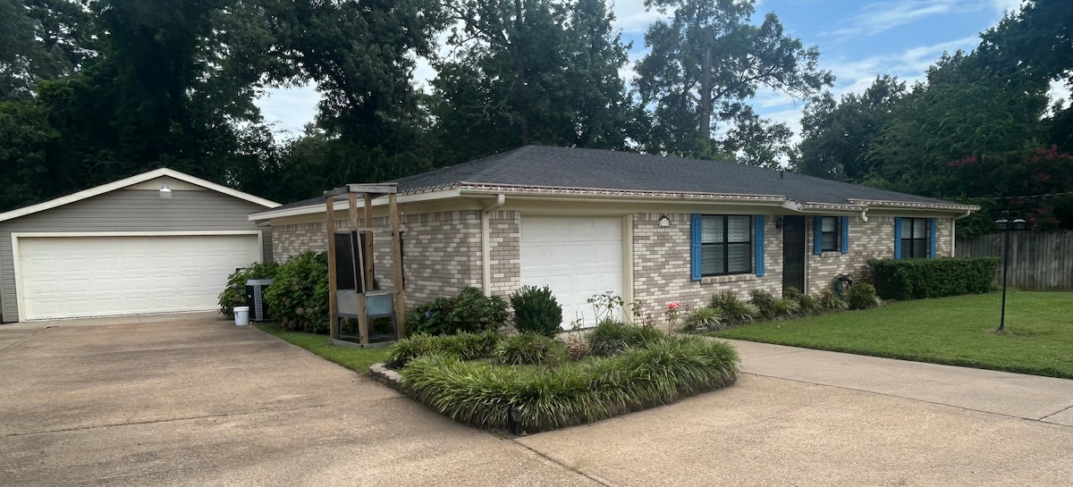 A charming single-story home is surrounded by lush greenery, with a neatly landscaped front yard. The brick facade is complemented by large windows that allow natural light. Two garage doors provide convenient parking space, and a driveway leads to the main entrance.