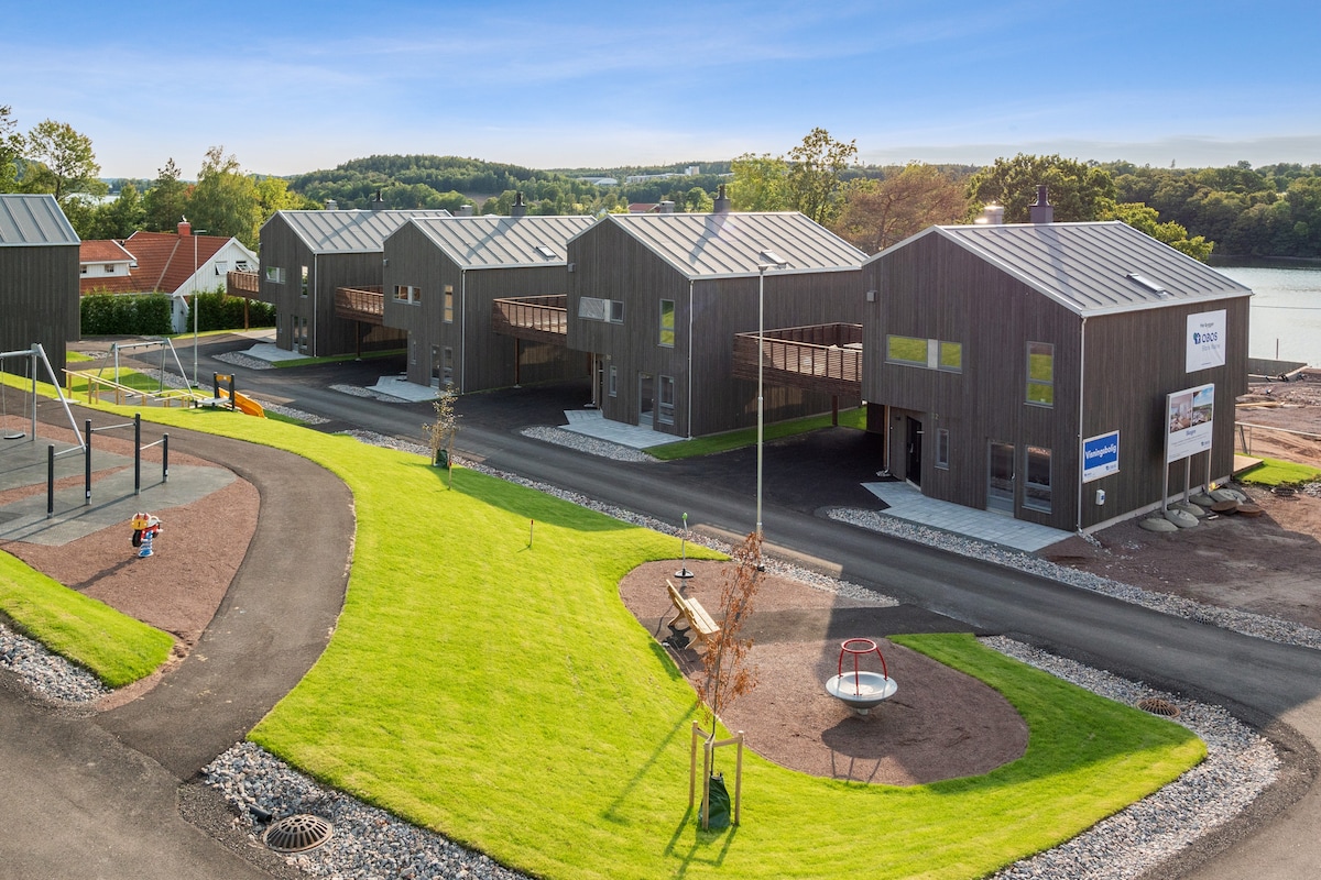 A modern property complex features multiple dark wooden structures with metal roofs. Green lawns surround the buildings, while a playground with swings and a roundabout is visible nearby. A walkway made of gravel leads past the units, providing access to the serene outdoor space.