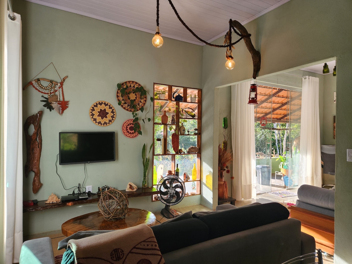 A living area features a dark sofa and a round wooden table, accented by decorative wall art and plant elements. A smart TV is mounted on the wall, with natural light streaming through large windows that open to an outdoor space.
