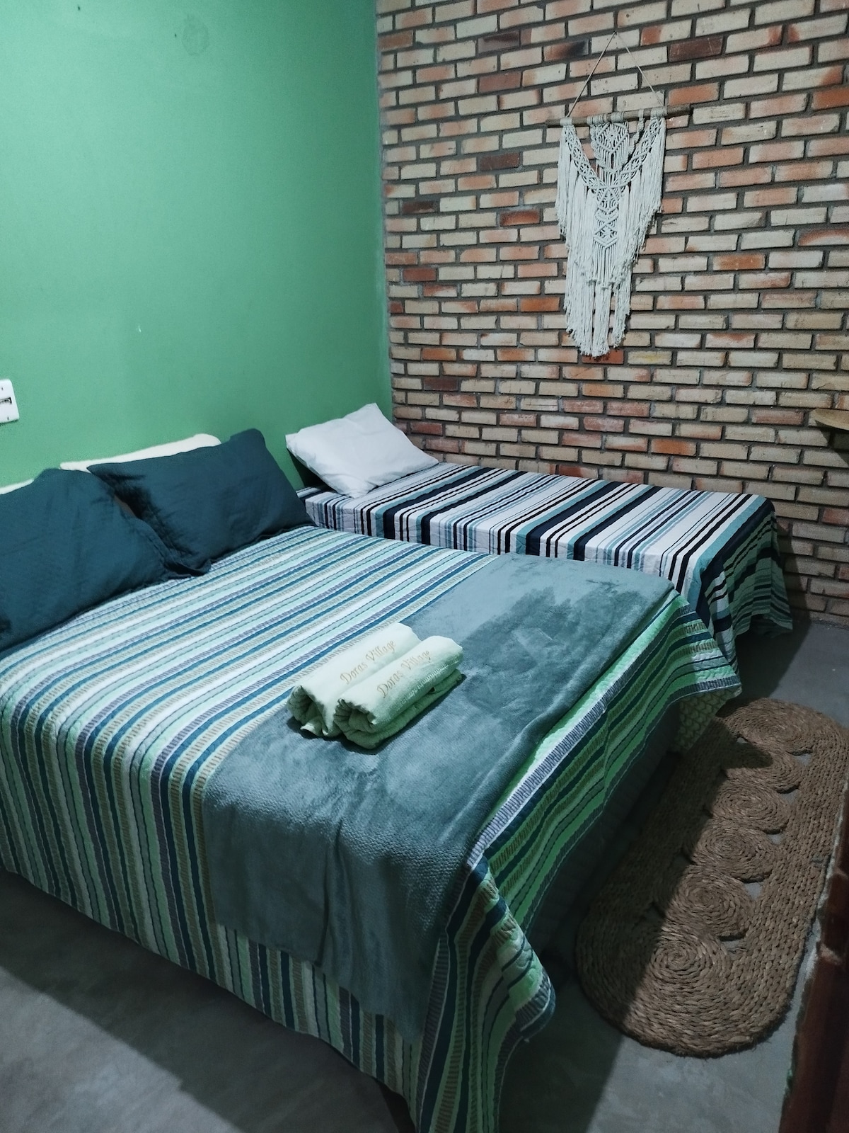 A sleeping area features two beds, one spacious and one narrower, each adorned with striped bed covers. Soft towels are neatly arranged at the foot of the beds, while a textured area rug adds warmth to the concrete floor. A patterned wall hanging complements the exposed brick backdrop.