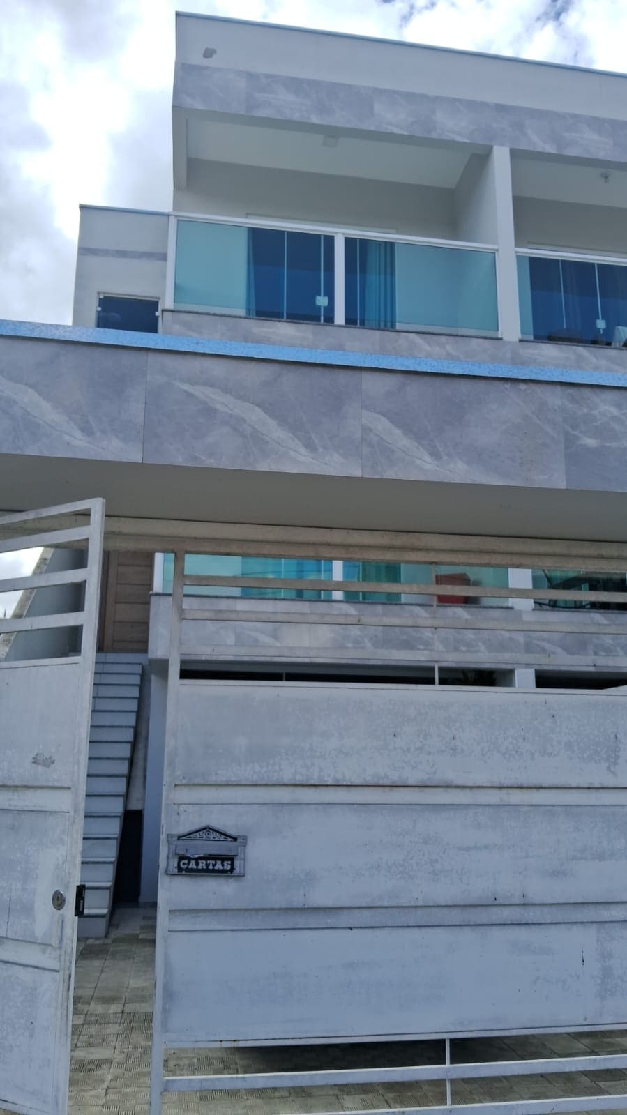 The exterior of a modern two-story building is depicted, featuring a combination of light grey walls and large glass balconies. An entrance gate is visible in the foreground, providing access to the property with staircases leading to the upper level.