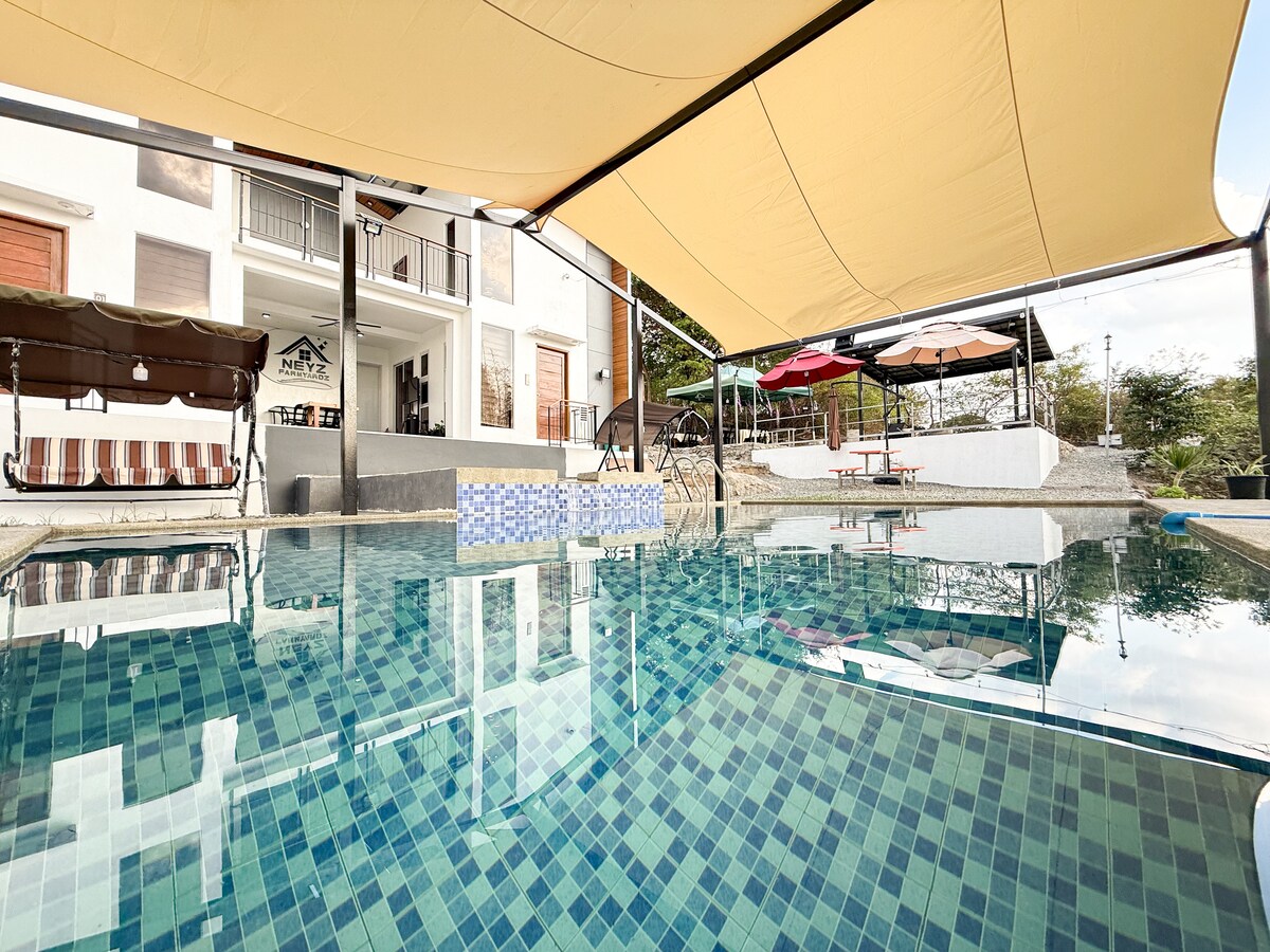 A scenic swimming pool showcases clear blue water surrounded by a patterned tile edge. Shade sails provide cover above, while various seating areas, including tents and tables, are visible in the background. The design promotes a relaxed outdoor atmosphere suitable for social gatherings.