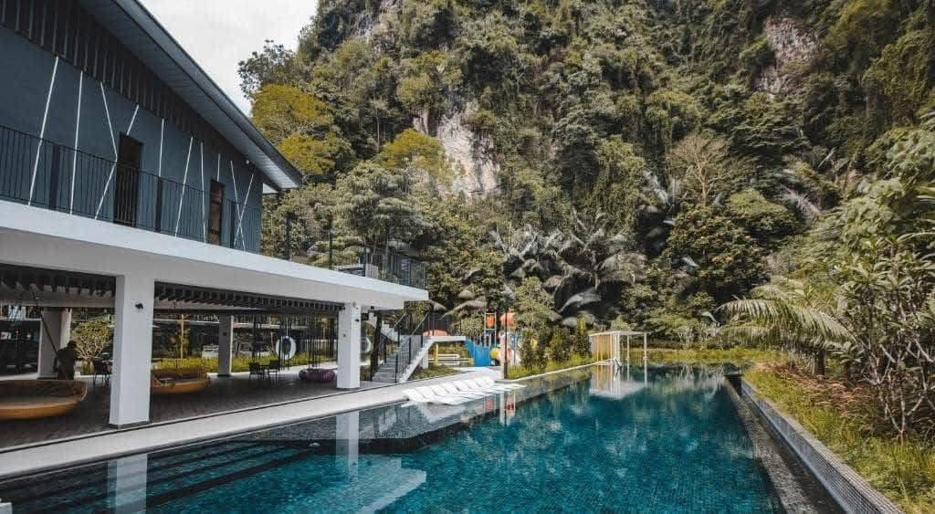 An expansive swimming pool is visible alongside a modern building, surrounded by lush greenery. The tranquil water reflects the nearby mountains, and colorful play structures can be seen in the background, indicating a family-friendly environment.