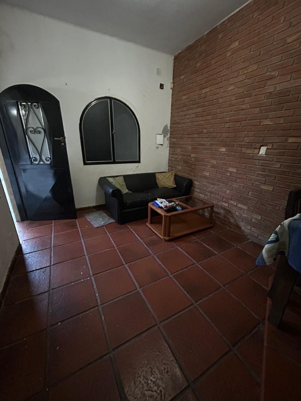 A simple living area is presented with a dark sofa positioned against a brick wall. A wooden coffee table sits in front of the sofa, containing various items. A doorway and window with an arched design provide access to natural light.