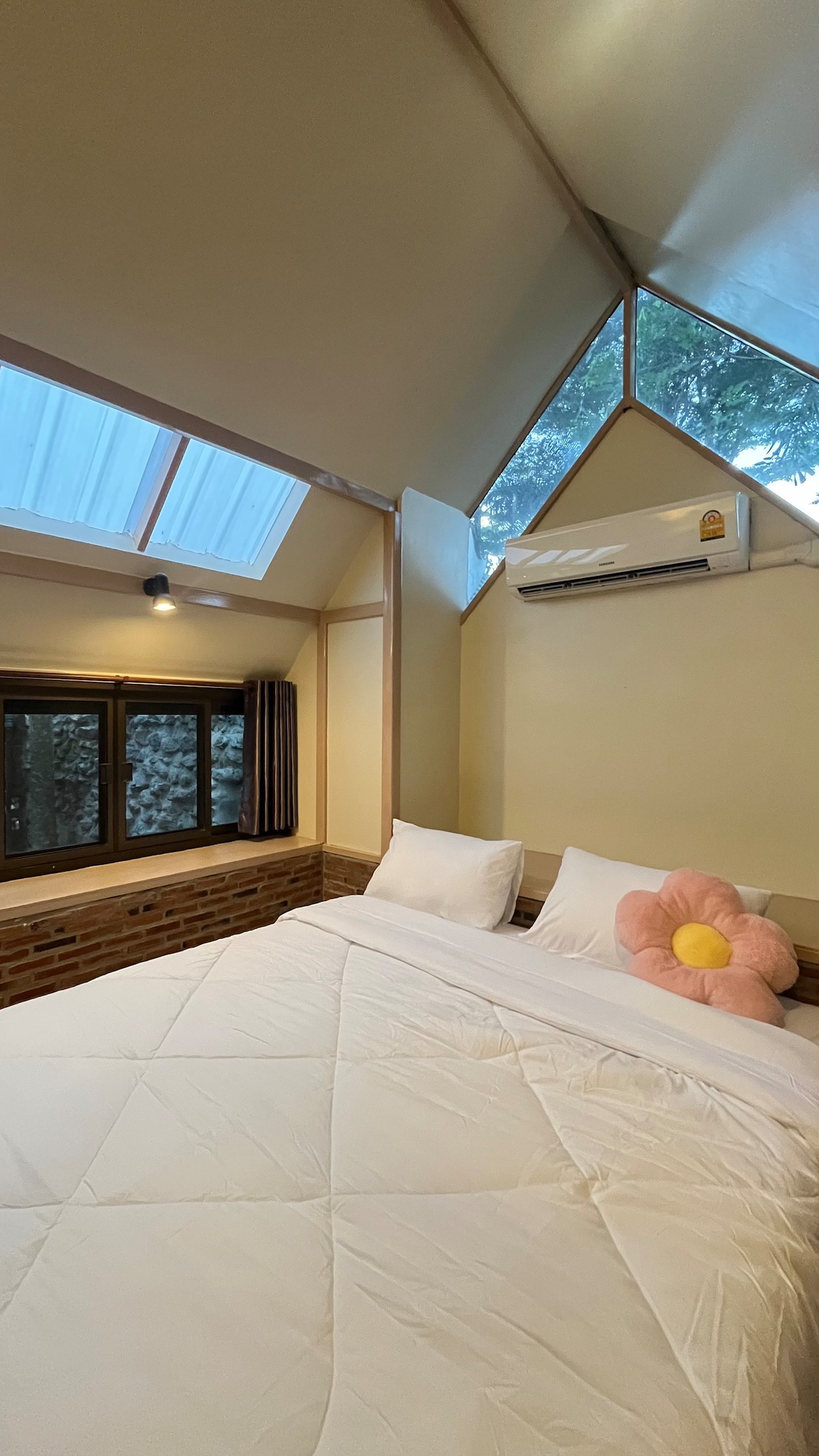A cozy room features a spacious bed, adorned with fluffy pillows and a large flower-shaped cushion. Natural light pours in through the sloped ceiling and windows, illuminating the warm, brick wall. An air conditioning unit is positioned above for comfort.