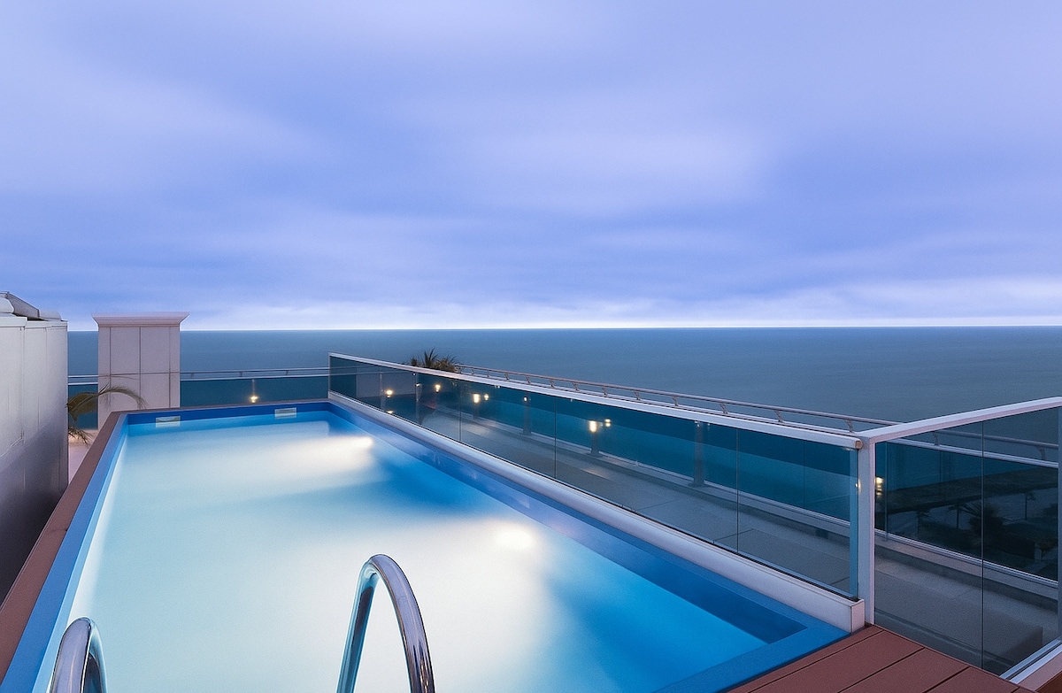 A rooftop pool is seen with crystal-clear water, framed by glass railings that provide unobstructed views of the sea. The calm evening sky creates a tranquil ambiance, enhancing the relaxation space designed for guests to enjoy.
