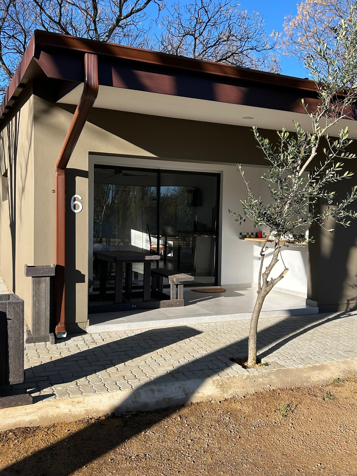 The exterior of a modern accommodation is presented, featuring a glass sliding door that opens onto a small porch. A simple wooden table with benches is visible inside. A slender tree stands beside the entrance, and the pathway is composed of stone pavers.