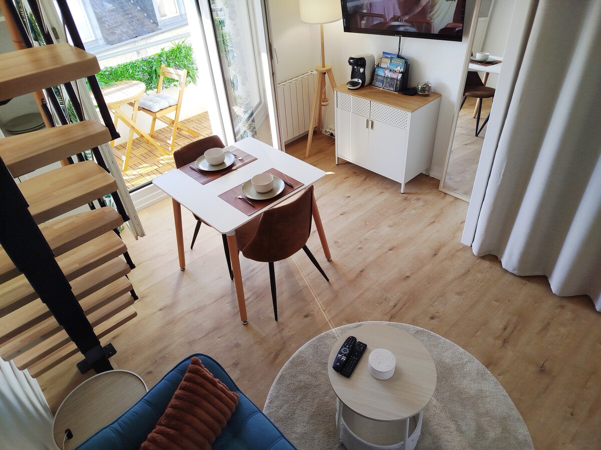 A cozy dining area features a small white table set for two, accompanied by two brown wooden chairs. A light-toned rug sits under a coffee table. A television is mounted on the wall, and a balcony with outdoor seating is visible through the glass doors.