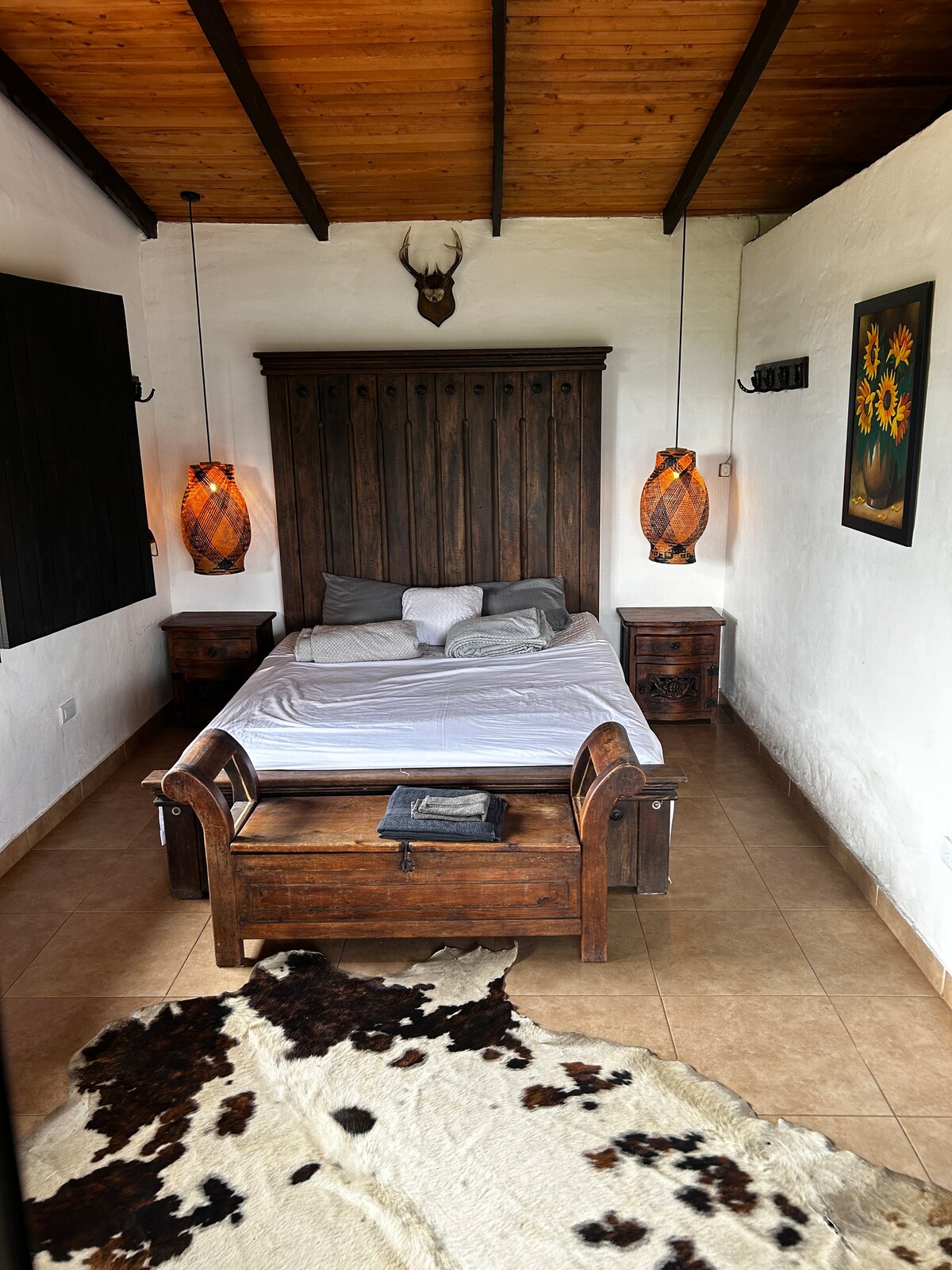 A cozy bedroom featuring a wooden headboard and a spacious bed dressed in neutral linens. Two woven pendant lights illuminate the room. Brown wooden nightstands are positioned on either side of the bed, and a decorative cowhide rug lies on the tiled floor.