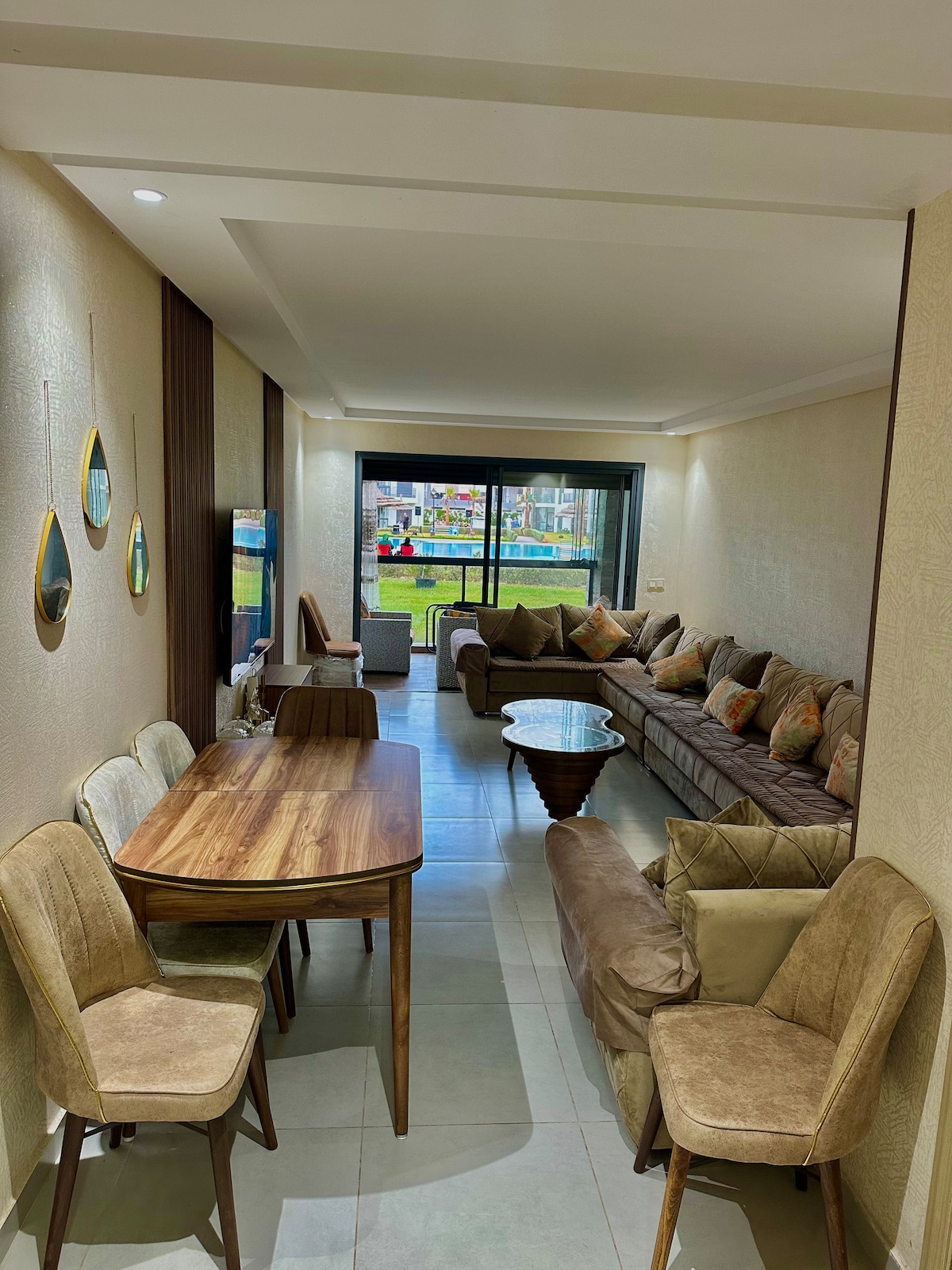 A light-filled living area is furnished with a spacious sectional sofa adorned with decorative pillows. A wooden dining table and six chairs provide seating for mealtime. Large sliding glass doors illuminate the space, revealing views of the outdoors.
