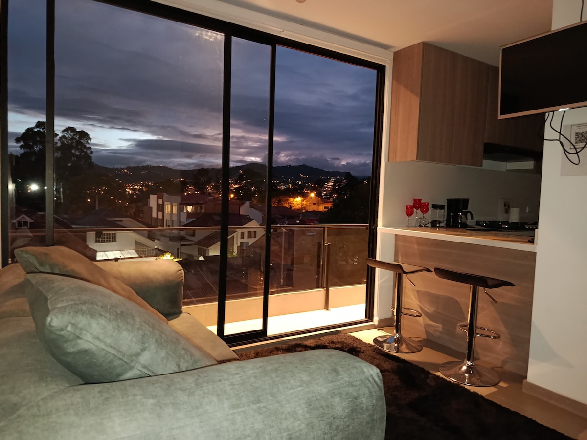 A modern living area features a comfortable sofa and two sleek bar stools positioned near a kitchen counter. Large glass doors invite natural light and showcase a city view at dusk, where warm lights twinkle in the distance. Dark carpet adds a touch of coziness.