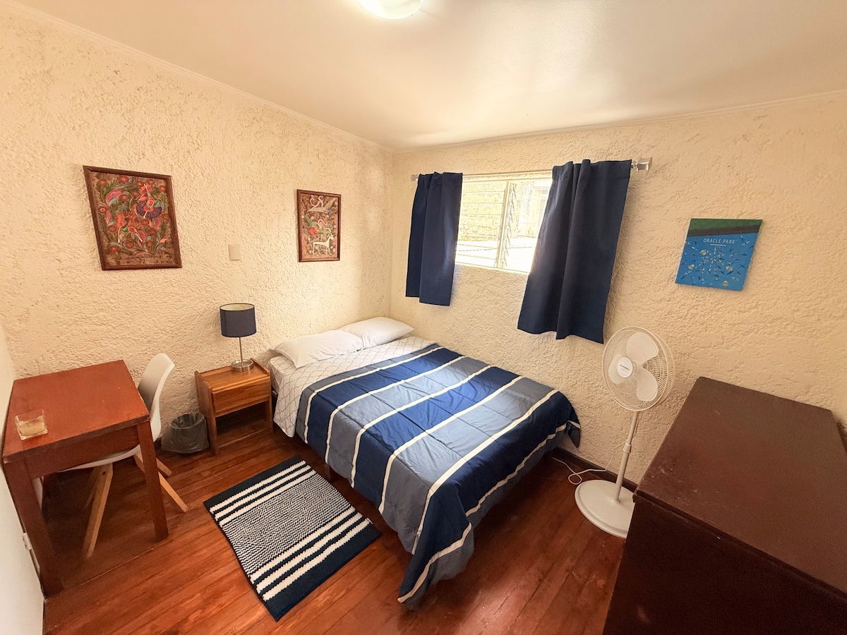 The room features a comfortable bed dressed in blue and white bedding. Two pieces of framed artwork are displayed on the textured walls. A small desk with a chair is positioned near a window covered by dark curtains. A wooden dresser and a standing fan are also visible.