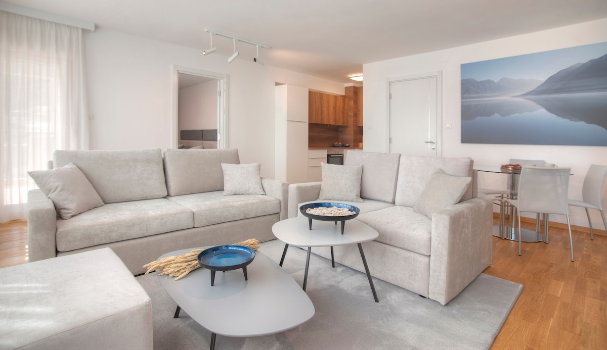A modern living area features a light-colored, plush sectional sofa and matching armchairs, arranged around two minimalist coffee tables. Natural light filters through sheer curtains, illuminating a tasteful wall art piece depicting a serene landscape. A dining area is visible in the background.