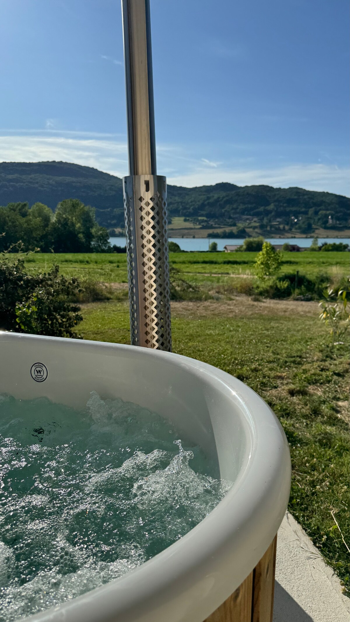 A close-up view of the nordic bath is shown, featuring bubbling water under clear skies. The tranquil landscape includes lush greenery and distant hills, with the Rhône River visible in the background, enhancing the serene setting.
